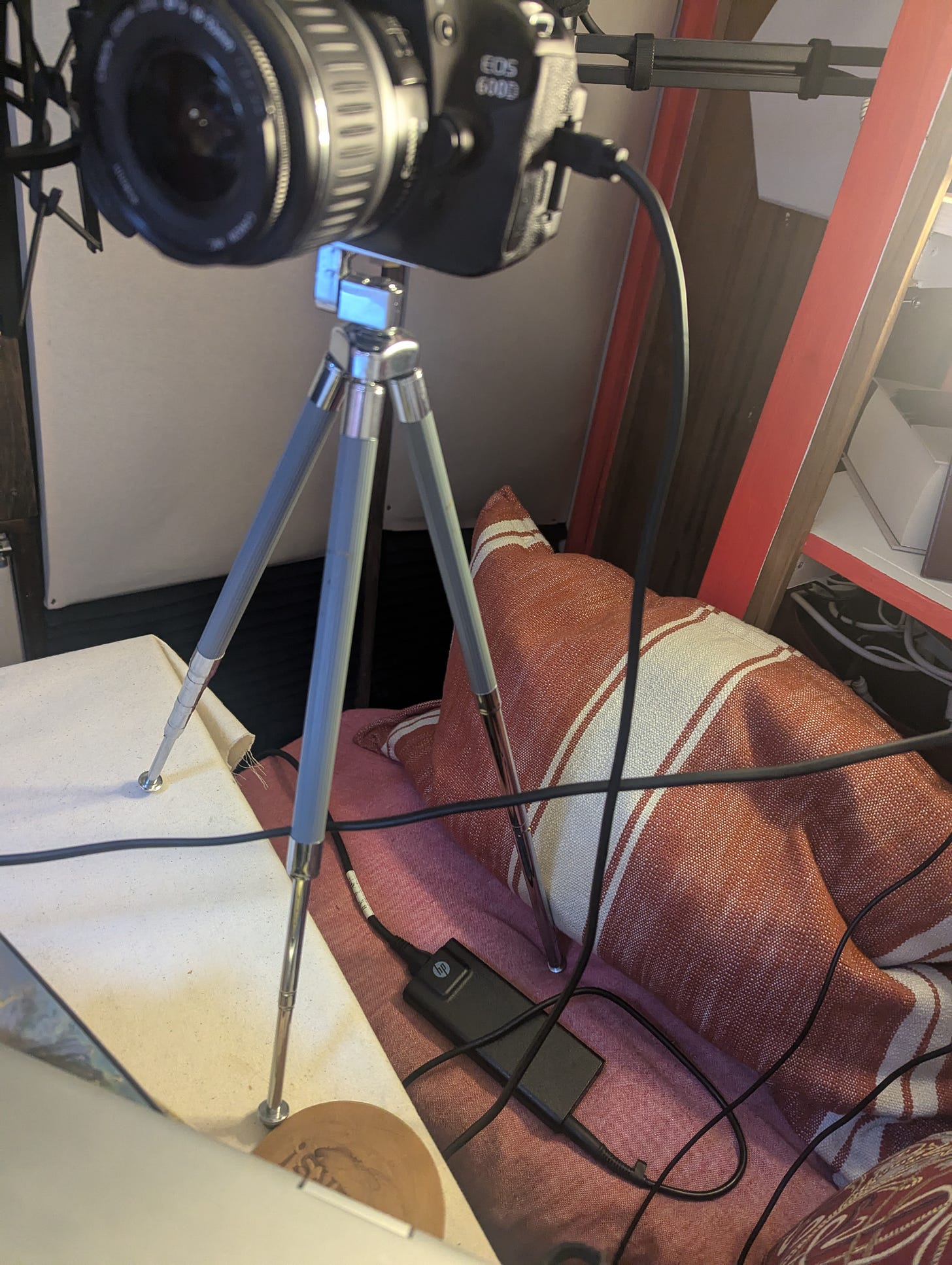 A tripod supporting a Canon camera. The camera is grey and silver. It is set up with two legs on a table, one on a chair.