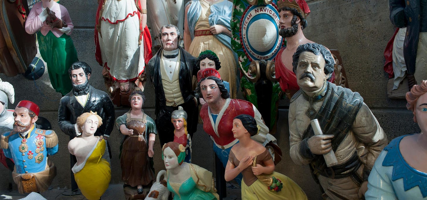 A photograph of a colourful grouping if 19th century ships' figureheads, men in suits, or women in dresses,, all looking out bravely or wistfully. They are all different sizes and the grouping is a surreal crowd