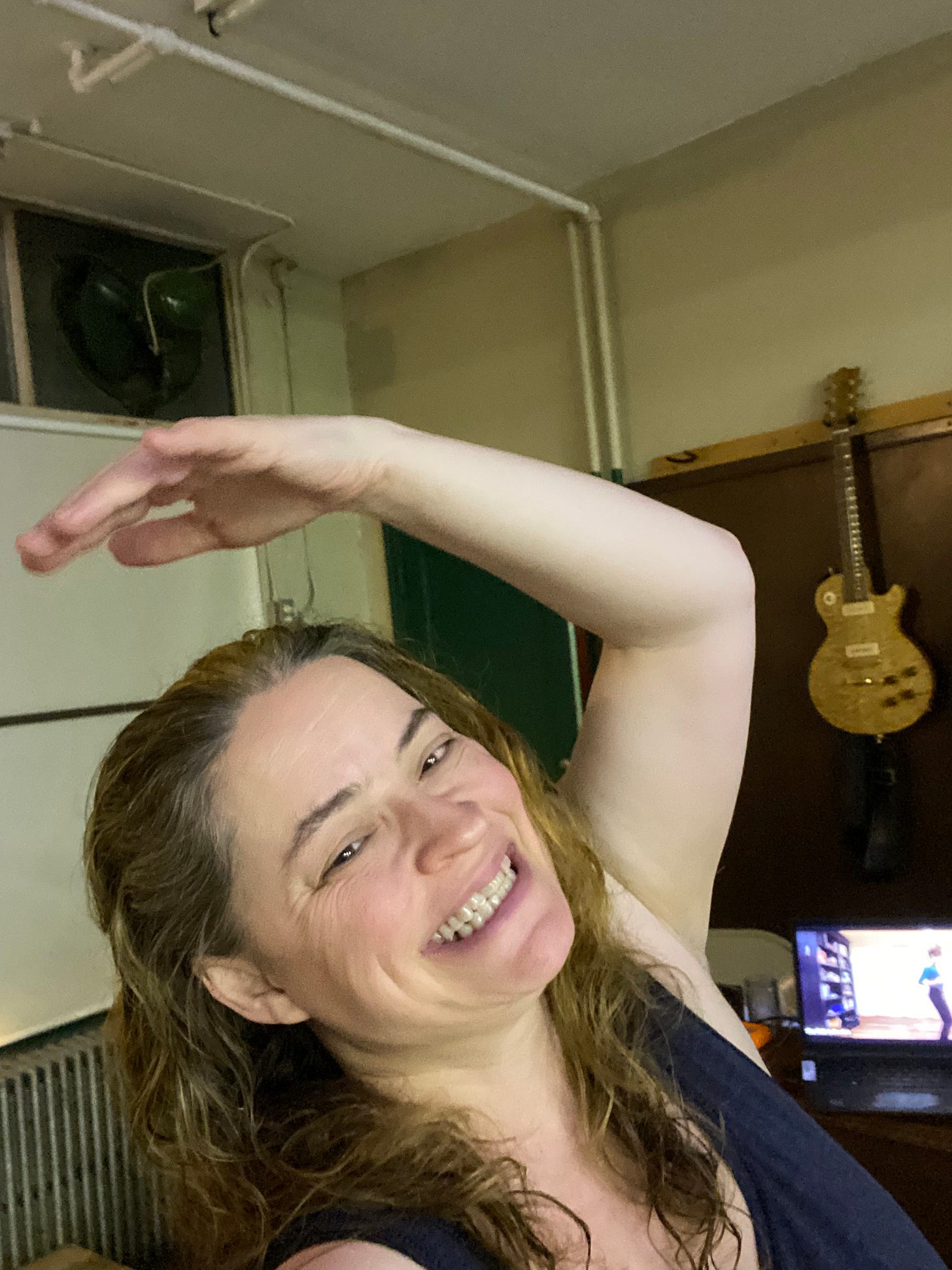 A femme presenting person smiling and lifting one arm in a studio space, with a dance class on the laptop behind them.