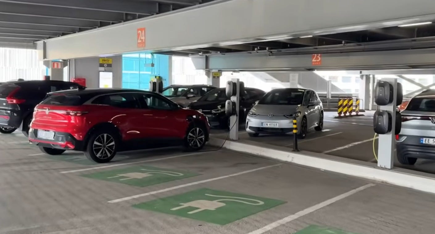 Video screenshot of a parking garage with six electric vehicles plugged into charging ports. All of the visible parking spaces are marked with  green paint with a sylized electric plug, indicating they're reserved for EV charging. (The video makes clear that there are three full rows of dedicated EV charging spaces, with additional rows of separate charging spaces in the garage for EV ride-sharing cars too)