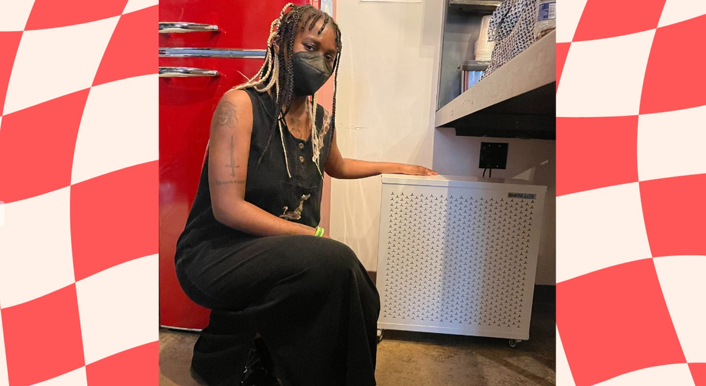 An organizer wearing a KN95 mask crouches next to an air purifier in a photo.