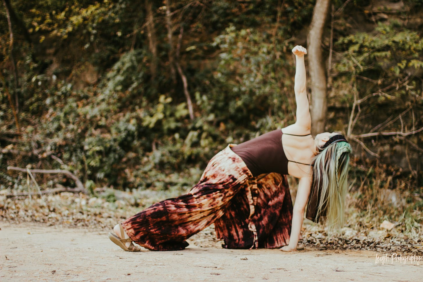The green-haired author wearing earth-tones in the woods: twisted into a deep lunge with one hand on the ground and other reaching for the sky.