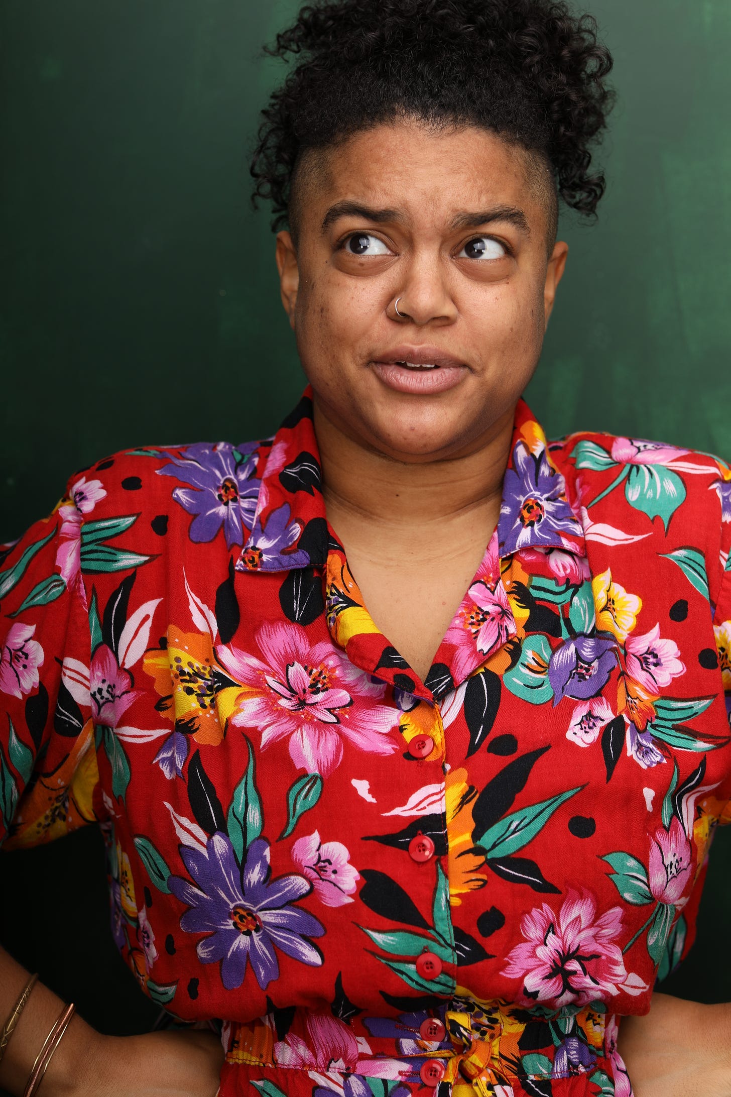 A photo of a light-skinned Black person with a silver nose ring looking to the left and making a silly face. They are wearing a colorful red floral top with buttons against a dark green background.