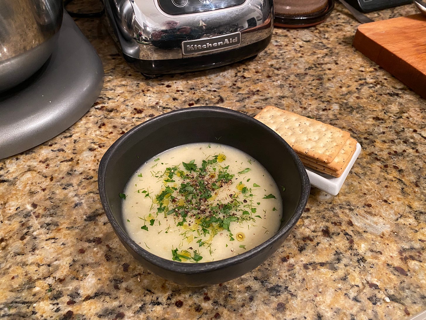 A small black bowl of potato leek soup, sprinkled generously with pepper, parsley, and dill, and drizzled lightly with olive oil. On a small white dish are a few rectangular crackers with poppyseeds in them.