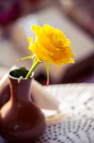 Picture of a single yellow rose in a small brown vase on top of a table.