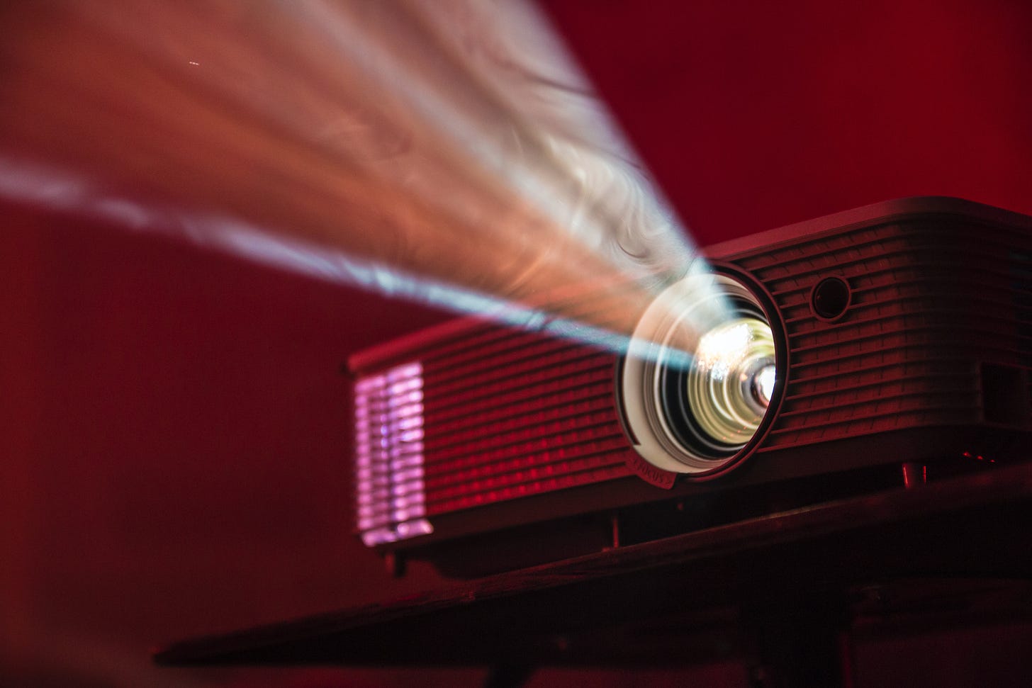 A moody image of an old slide projector, with red and purple hues. The projector is on and the light is streaming out from the machine.
