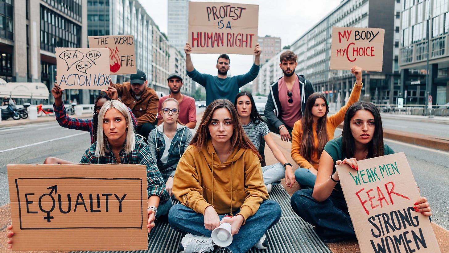 Group of young women and men sat on a road in a city. Holding up feminist protest signs.