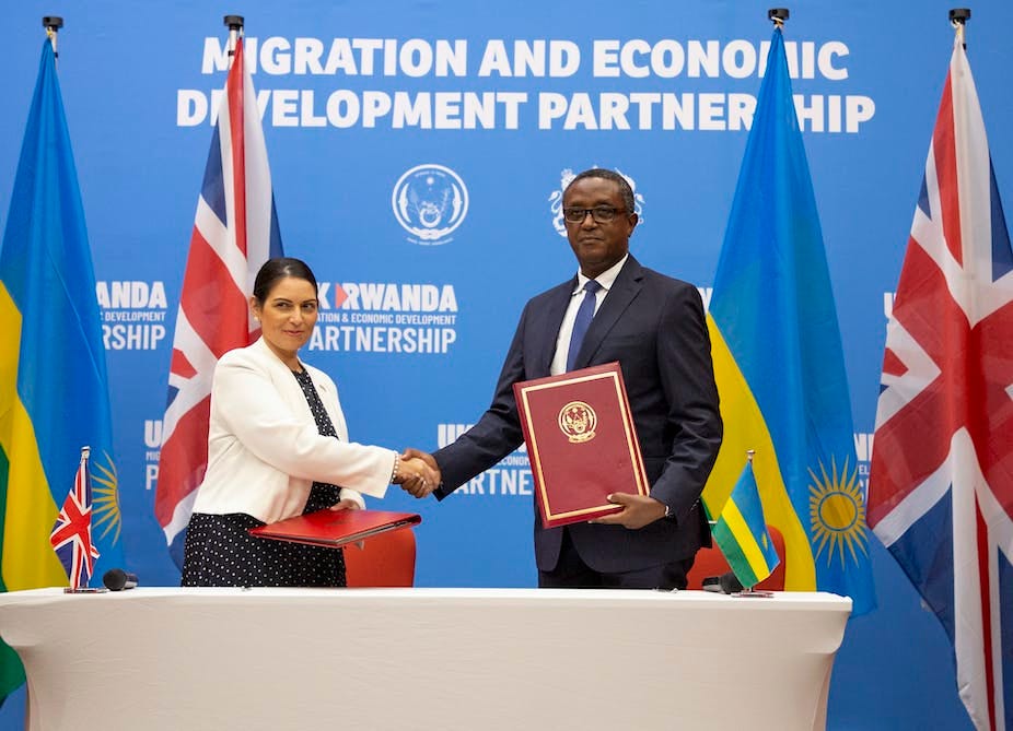 British Home Secretary Priti Patel (L) with Rwandan Foreign Minister Vincent Biruta shake hands in front of a blue wall reading Migration and Economic Development Partnership, and four Rwandan and UK flags.