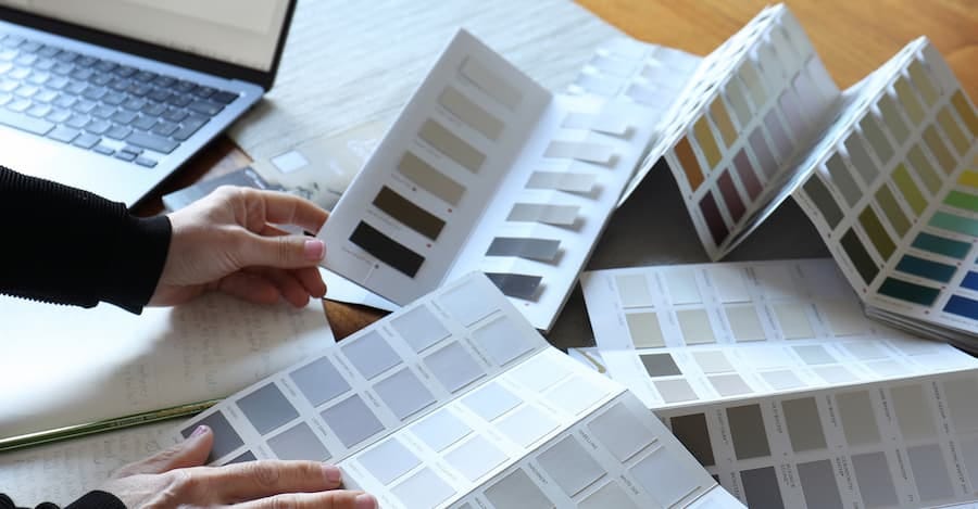 A person’s hands holding open a colour swatch card. The card is open on a page with different shades of grey. There are other colour swatch cards scattered around the table. There is a notebook and laptop on the table.