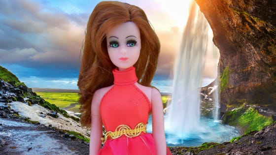 A delicate Dawn Doll with green eyes and long red hair stands before a pounding waterfall with idyllic puffy clouds overhead.