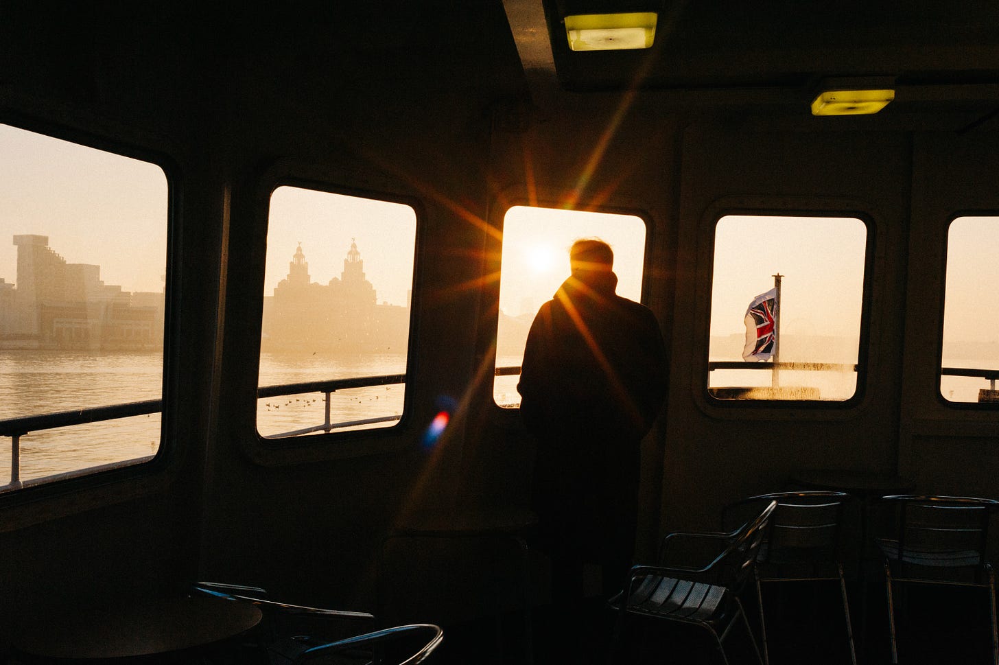 The Mersey Ferry crosses the river at sunrise. The sun is a giant star shining through the window of the ferry. A man looks out the window towards the waterfront.