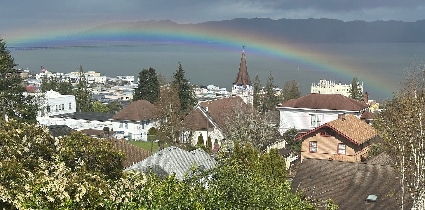 A rainbow is visible arcing over the waterfront of the small town of Astoria, Oregon. Rooftops of buildings in the town are visible in the foreground. The columbias river cuts through the middle of the image with a bridge visible in the distance and mountains shadowed by cloud in the background on the other side of the river.