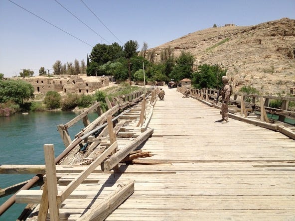 This image of a bridge taken from one end looking along its length is in Kajaki, Afghanistan (north central). A group of Marines in military gear crossing a damaged wooden bridge over a body of water in a dry, mountainous region. The bridge has broken and missing planks, along with makeshift wooden railings, indicating wear or conflict-related damage. In the background, there are mud-brick buildings, green vegetation, and hills.  One Marine is standing guard on the right side of the bridge midway across, holding a rifle, while others are walking further ahead. The sky is clear and bright during a sunny day.