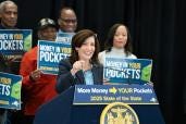 Governor Kathy Hochul standing at a podium with a microphone, announcing New York State's first-ever Inflation Refund proposal, with a group of people including Micah Shrewsberry, M. M. Mani, and Chanda Rubin in the background.