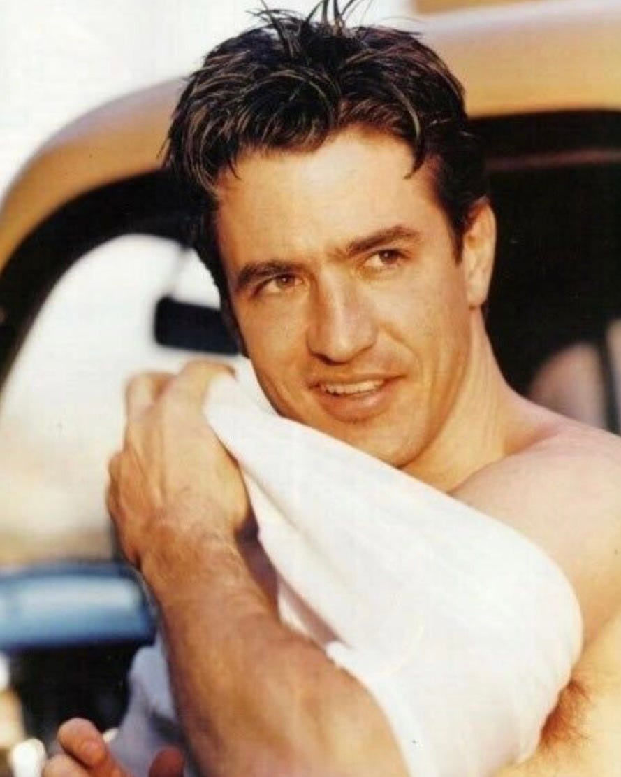 An image of Dermot Mulroney from the '90s. He has his arm through one arm hole of a white t-shirt as he puts the shirt on. He looks off to the side with a crooked smile.