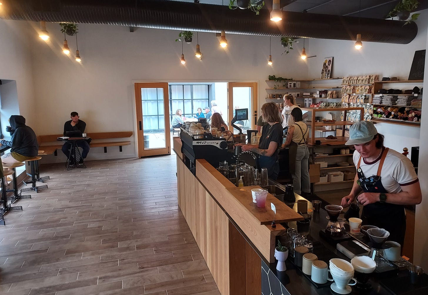 A wide shot of a coffee bar and open coffee shop space leading to an outdoor patio. On the right baristas pull shots at an expresso machine and make pourover style coffees. The coffee bar is a soft blonde wood.