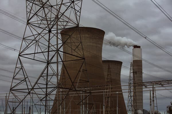 White smoke pours from a brown and white smokestack next to two large cooling towers. Power lines surround the structures. 