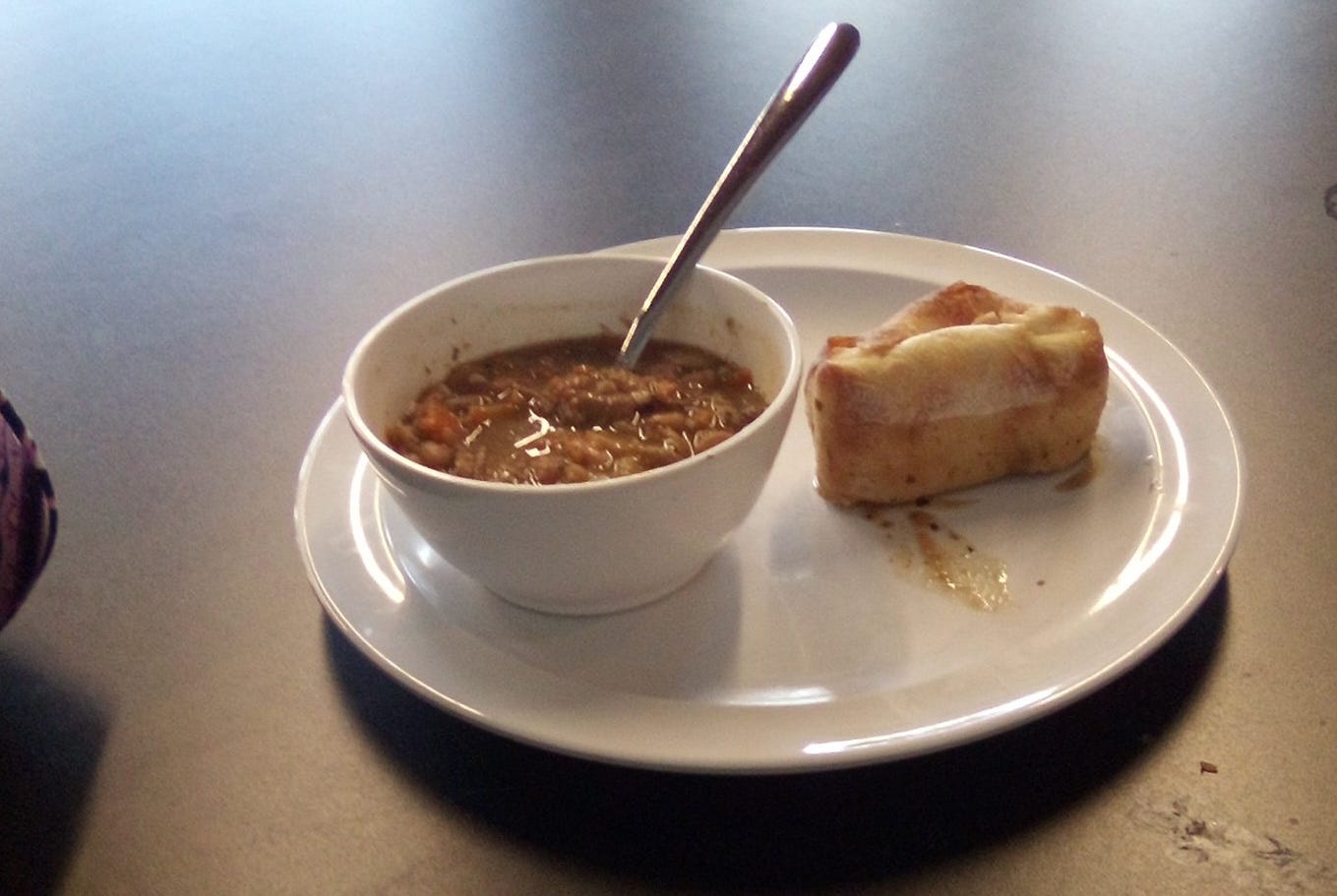 Image of a small white bowl of ham and bean soup, with a spoon inserted, on a plate with a piece of bread, all set on a metallic table