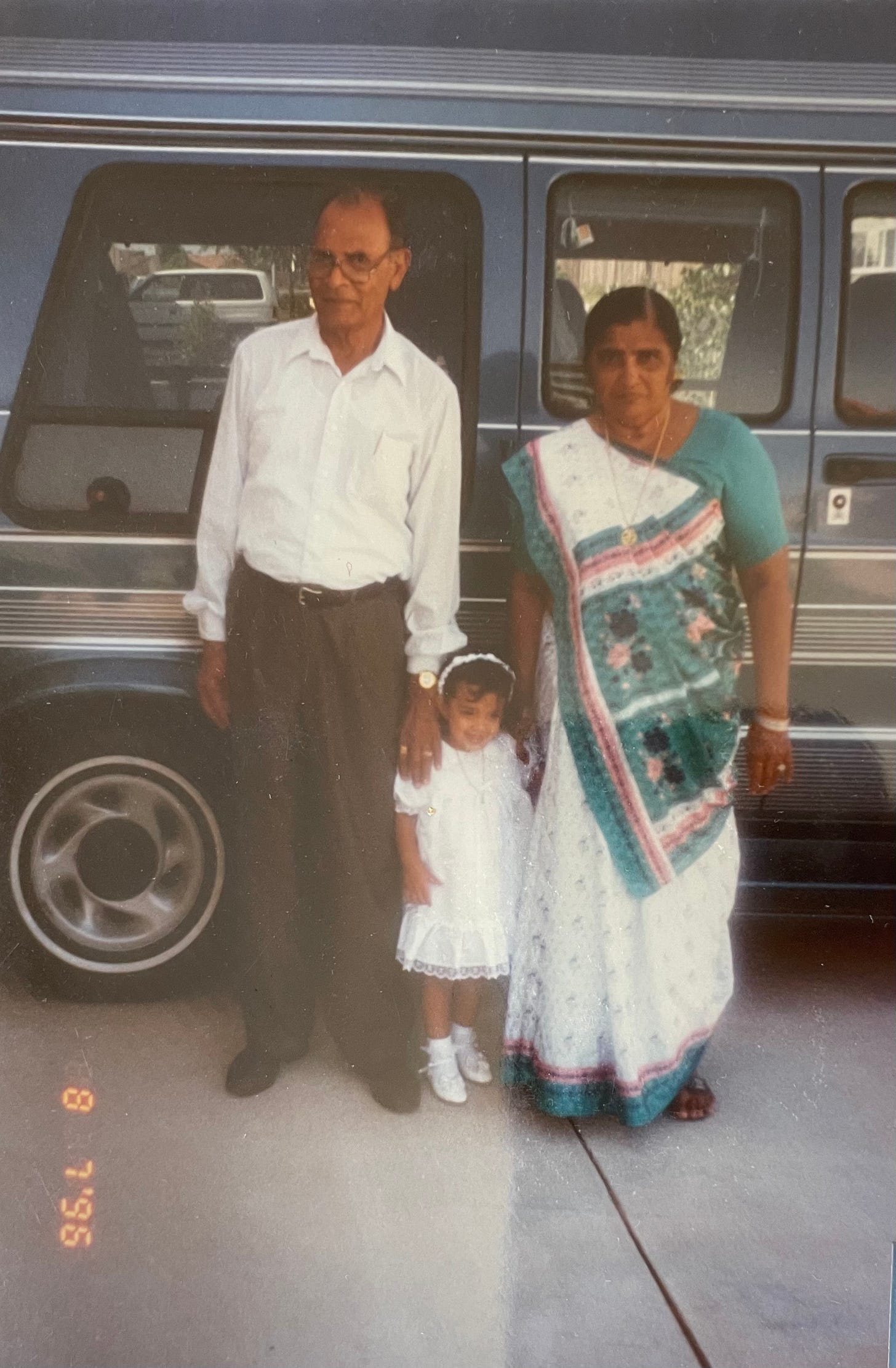 An elderly man wears a white shirt and dark dress pants. He stands with his left hand on a toddler's shoulder, who wears a white dress. A woman, right, wears a white, pink, and green saree, with her right hand on the toddler's shoulder. They stand in front of a blue van.