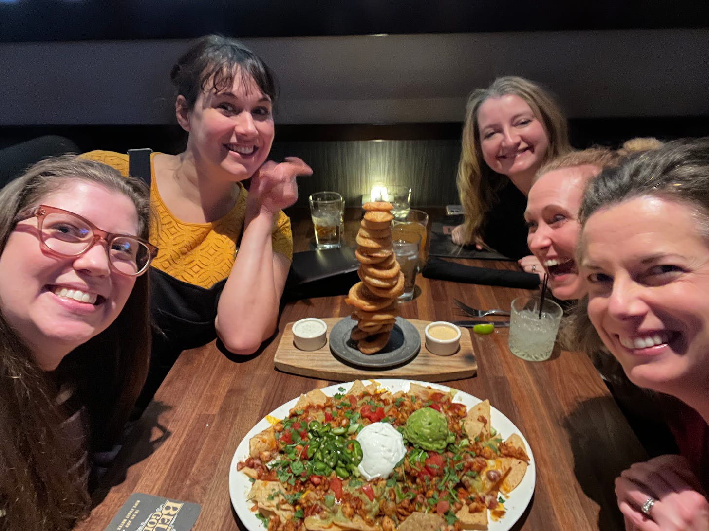 5 smiling women around a table; on the table is a giant stack of onion rings and a huge plate of nachos