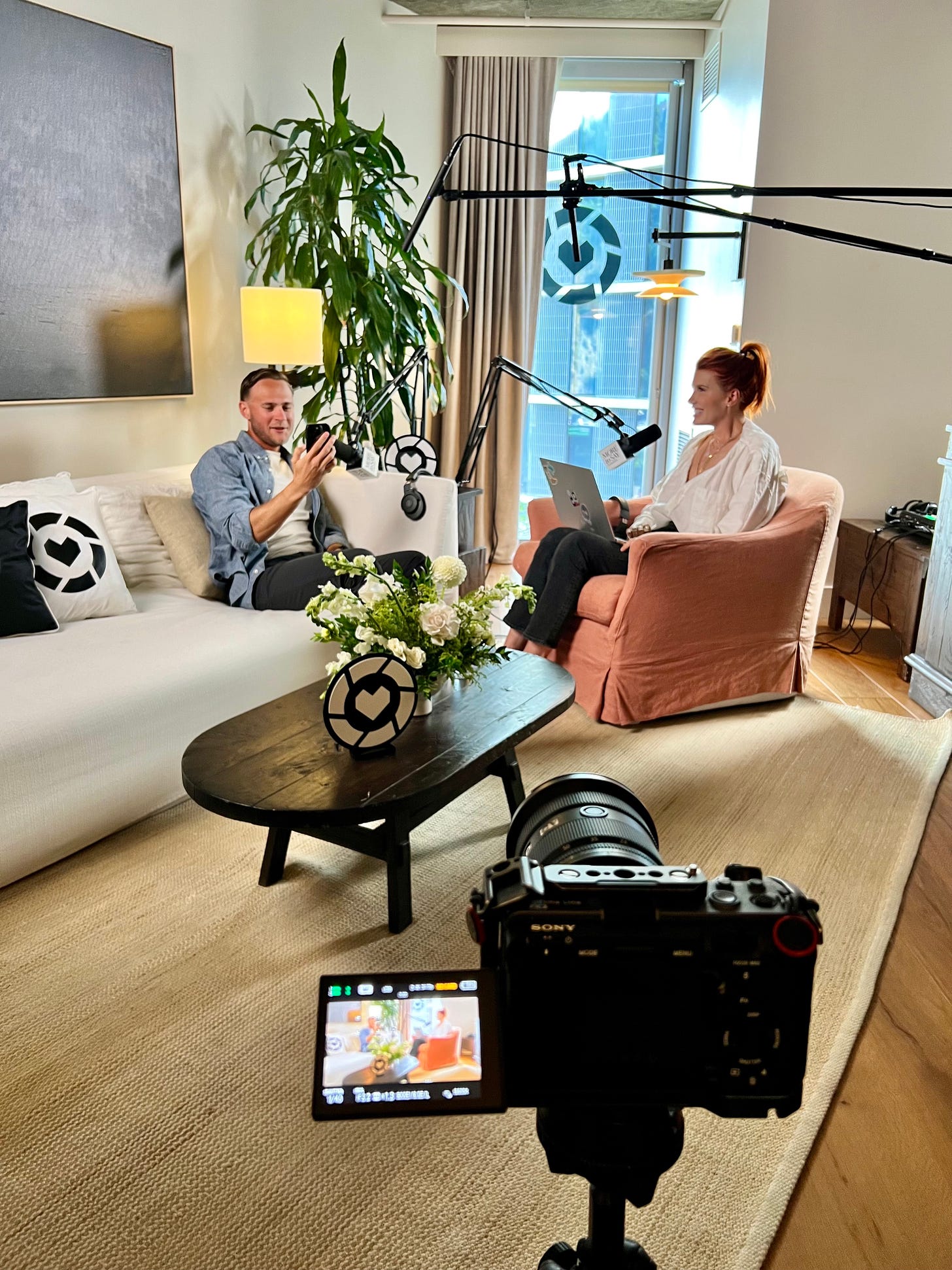 Image of a suite with Paul Johnson and Amber Venz Box recording a podcast called More to Say. LTK branding is on the pillows, mic, windows and decorative elements on the coffee table between them