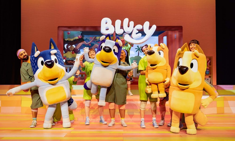 Image from Bluey's Big Play showing Bluey and her family in large puppet form with visible puppeteers. Bluey