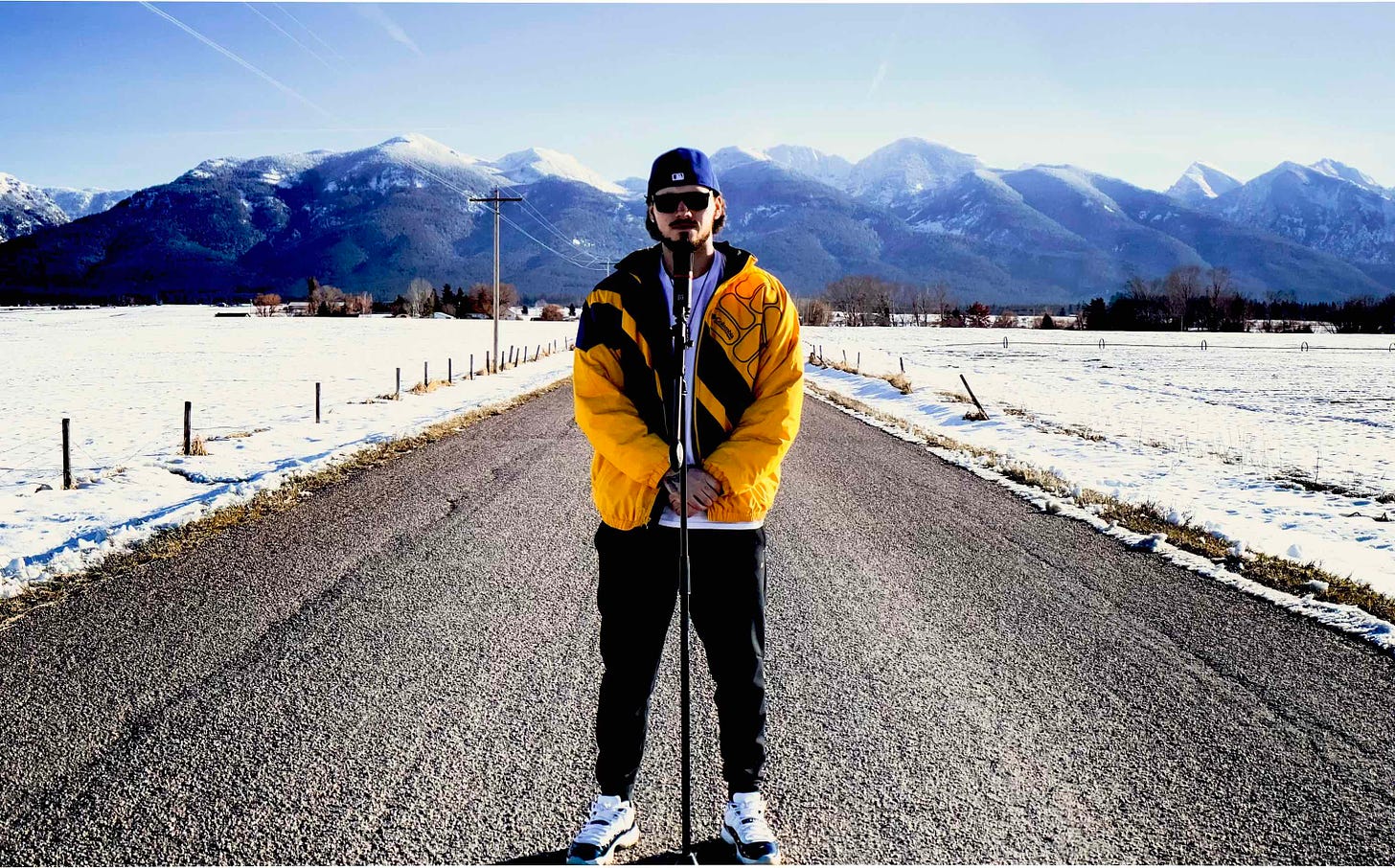 man in shades yellow jacket with mic on snowy road with mtns in background