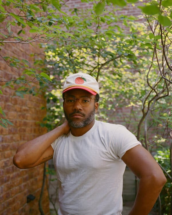 A man with a lightly graying beard, wearing brown glasses and a ball cap with an orange on it, poses outside in a white T-shirt, bringing his right hand up to grab the back of his neck.