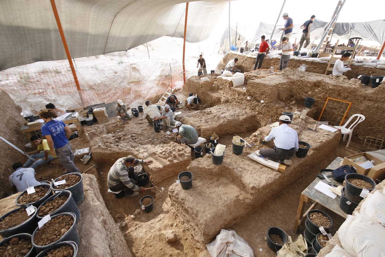 Archaeologists working on a large, terraced excavation area under a white canopy.