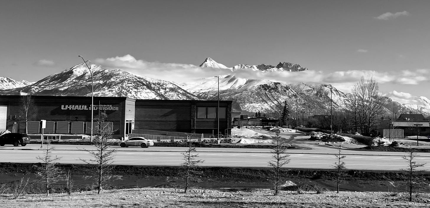 Black and white picture of snow-dusted mountains in the background, a U-haul store in the mid-ground, and a highway in the foreground