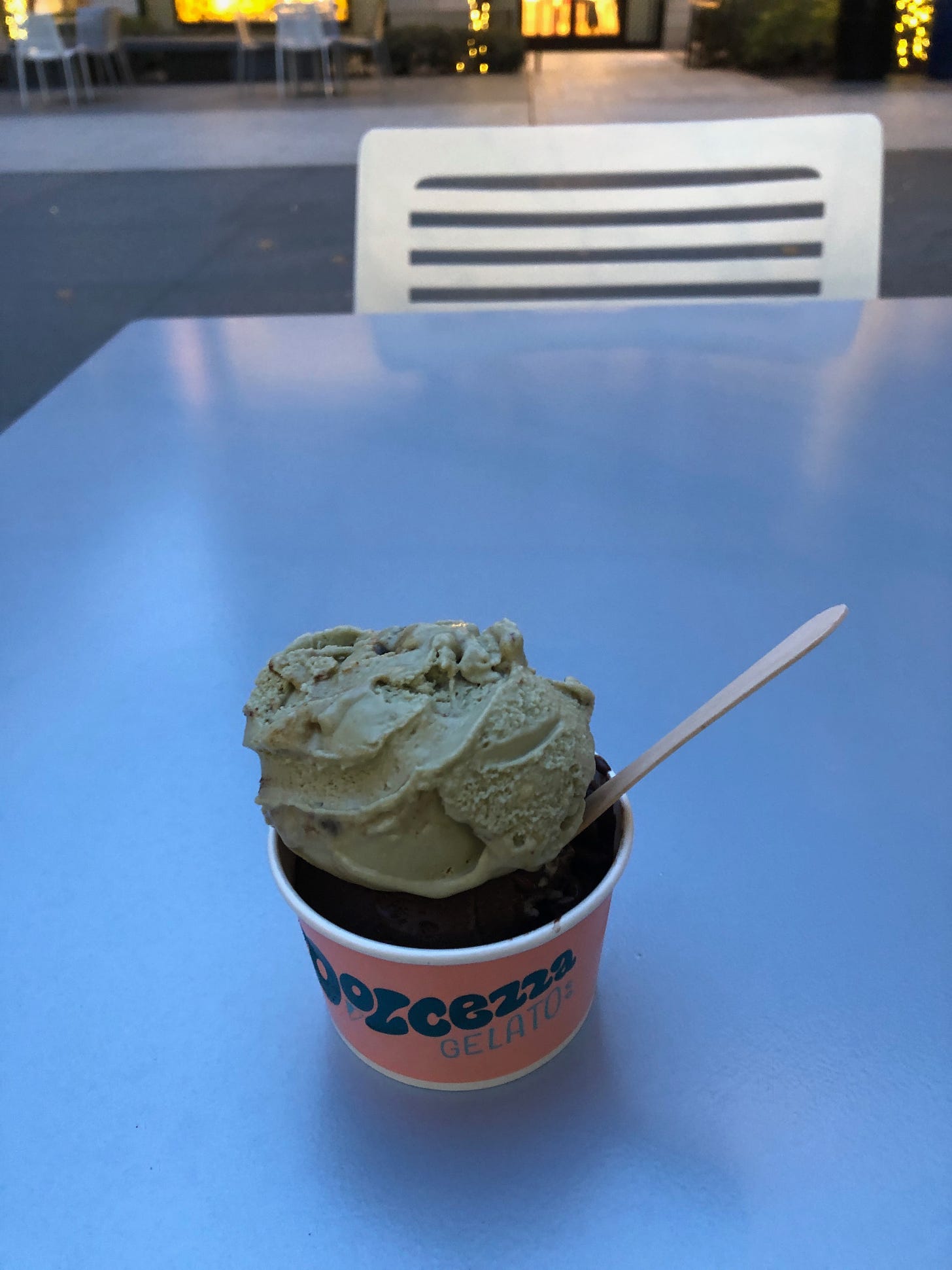 Two scoops of ice cream in a cup topped by a wooden spoon. The cup is on a small table facing a chair. In the background are brightly-lit stores and more outdoor tables and chairs.