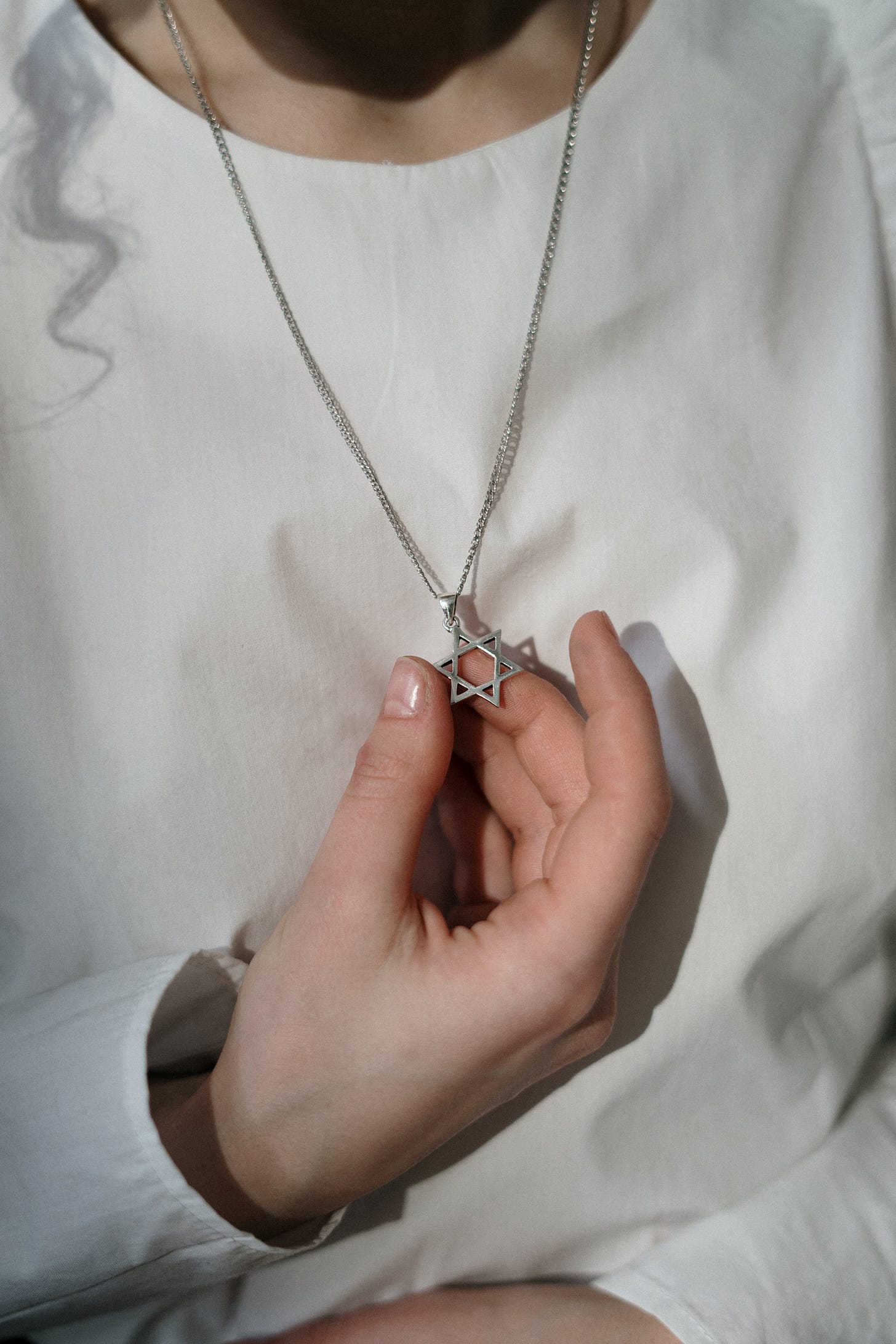 Woman's hand holding a Jewish star necklace against her white shirt, with the shadow of a lock of curly hair.
