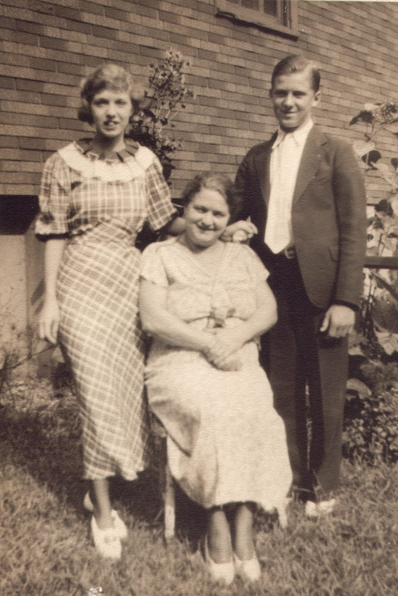 Young woman in plad dress, older woman seated, young man in dark suit and white dress shirt.