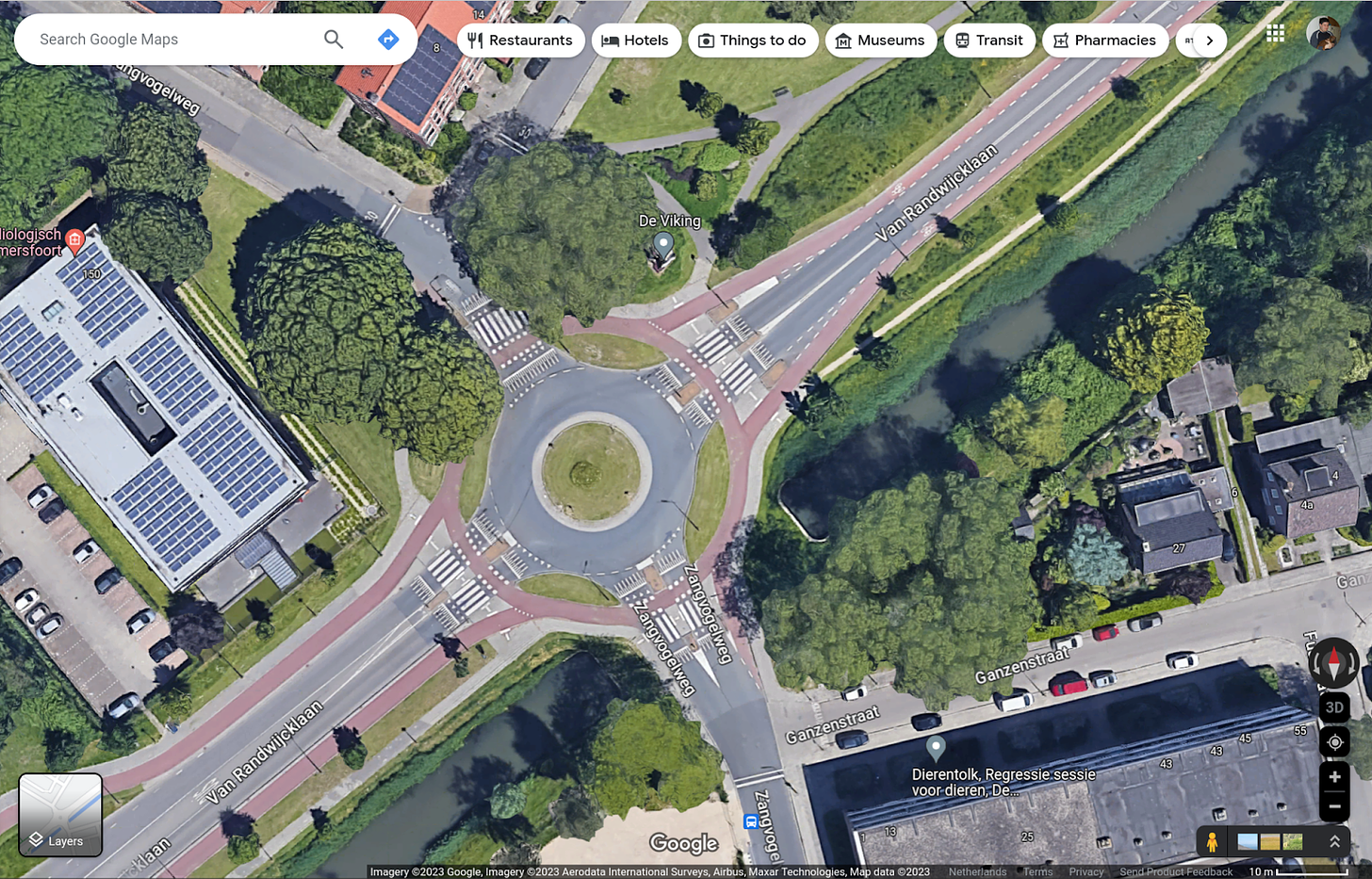 A Google maps satellite view screenshot showing a large traffic circle with a red protected cycleway around it. To the right, the red cycleway becomes red on-street bike lanes and to the left, it continues as a protected cycleway.