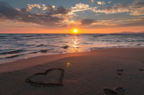 Free Beautiful sunset over ocean with a heart shape in the sand, perfect for romantic themes. Stock Photo