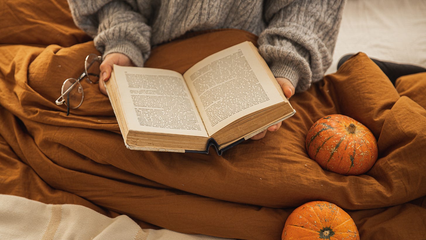Stock photo of white hands holding an open book, the person wears a grey sweater and sits under a mustard-coloured blanket. Pumpkins are placed on top of the blanket.