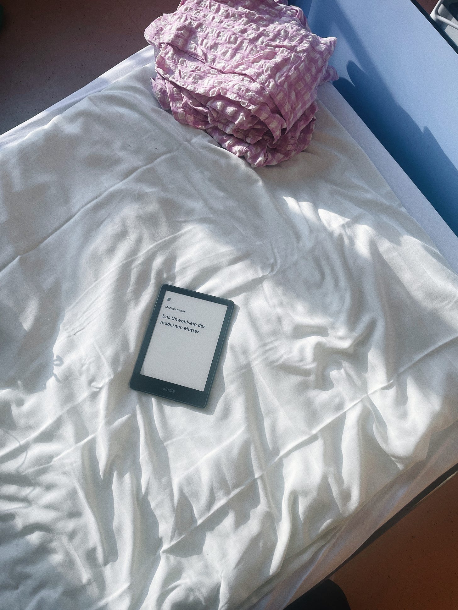 Auf dem Bild sieht man ein gemachtes Krankenhausbett mit weißer Bettwäsche. Das Licht fällt hart seitlich durch ein Fenster darauf. Auf dem Bett liegen ein gefalteter, rosa-weiß-karierter Pyjama sowie ein angeschalteter Kindle. Auf dem Kindle ist der Buchtitel „Das Unwohlsein der modernen Mutter“ von Mareice Kaiser zu sehen.