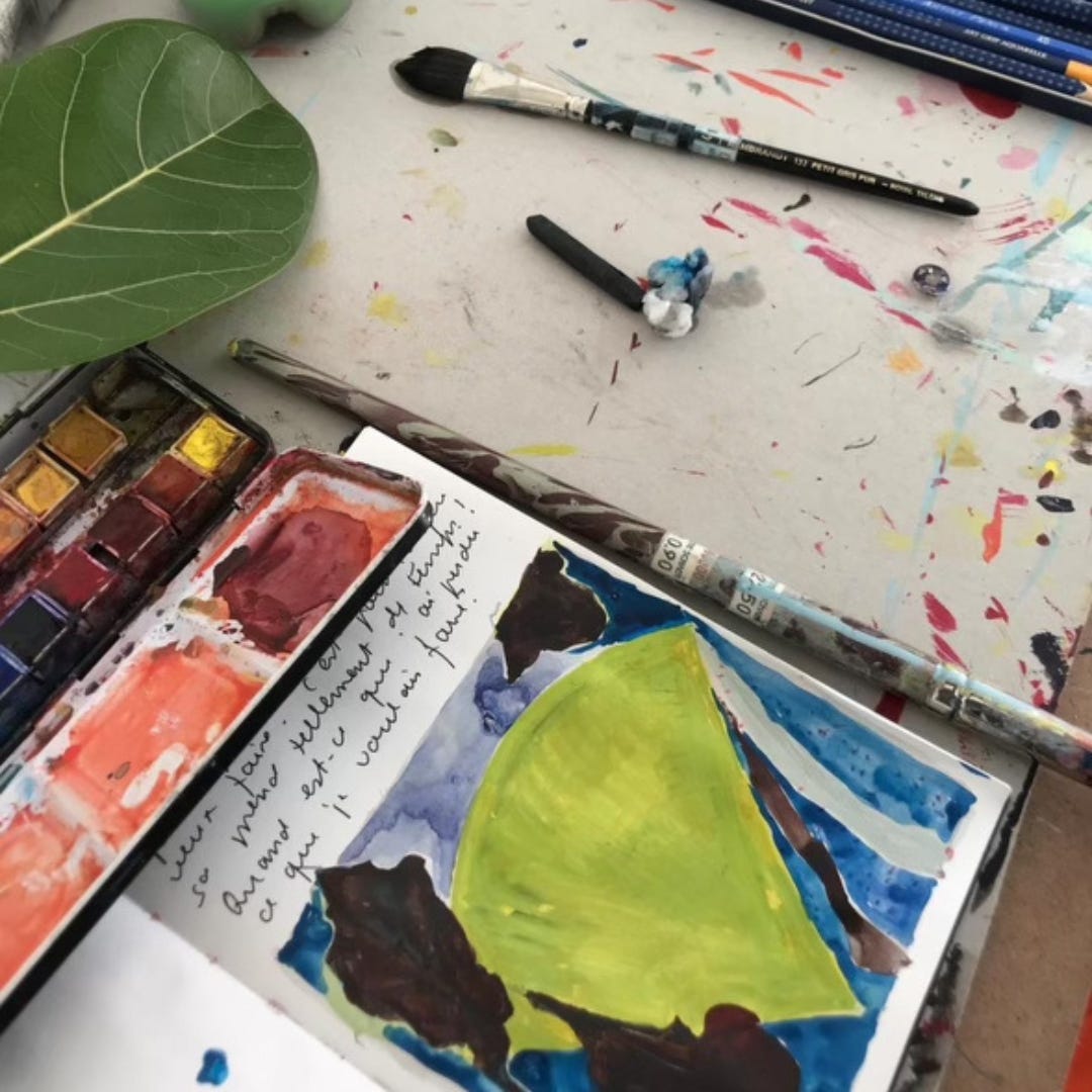 Photograph of a tabletop that contains an open journal, the page split between handwritten text in French and an abstract watercolor painting rendered in blues, purples and greens. The journal rests on a tabletop marked with smudges of paint and ink. Around the journal is a heavily used watercolor set, a scattering of paintbrushes and pens and the smooth green leaf of a fiddle leaf fig tree.