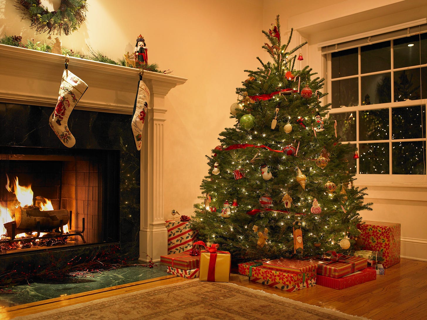 A Christmas tableau of a cosy home with a beautifully directed Christmas tree, under which are many wrapped presents. To the left of the tree, a warm fire, stockings hanging from the mantle.