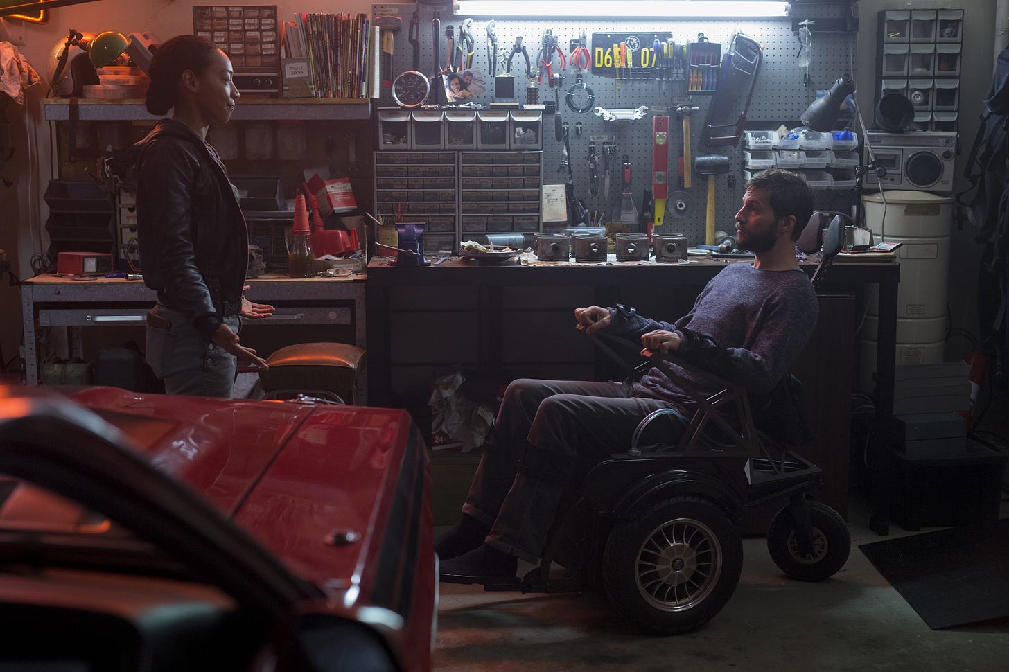 A cluttered garage with plenty of old-school tools all around. A woman standing on the left, facing a man in the wheelchair. They appear to be talking.