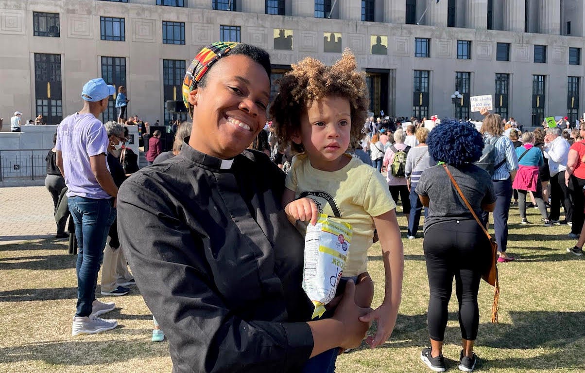 Jen at the rally outside the State House, wearing a clerical collar and holding her 2 year old son
