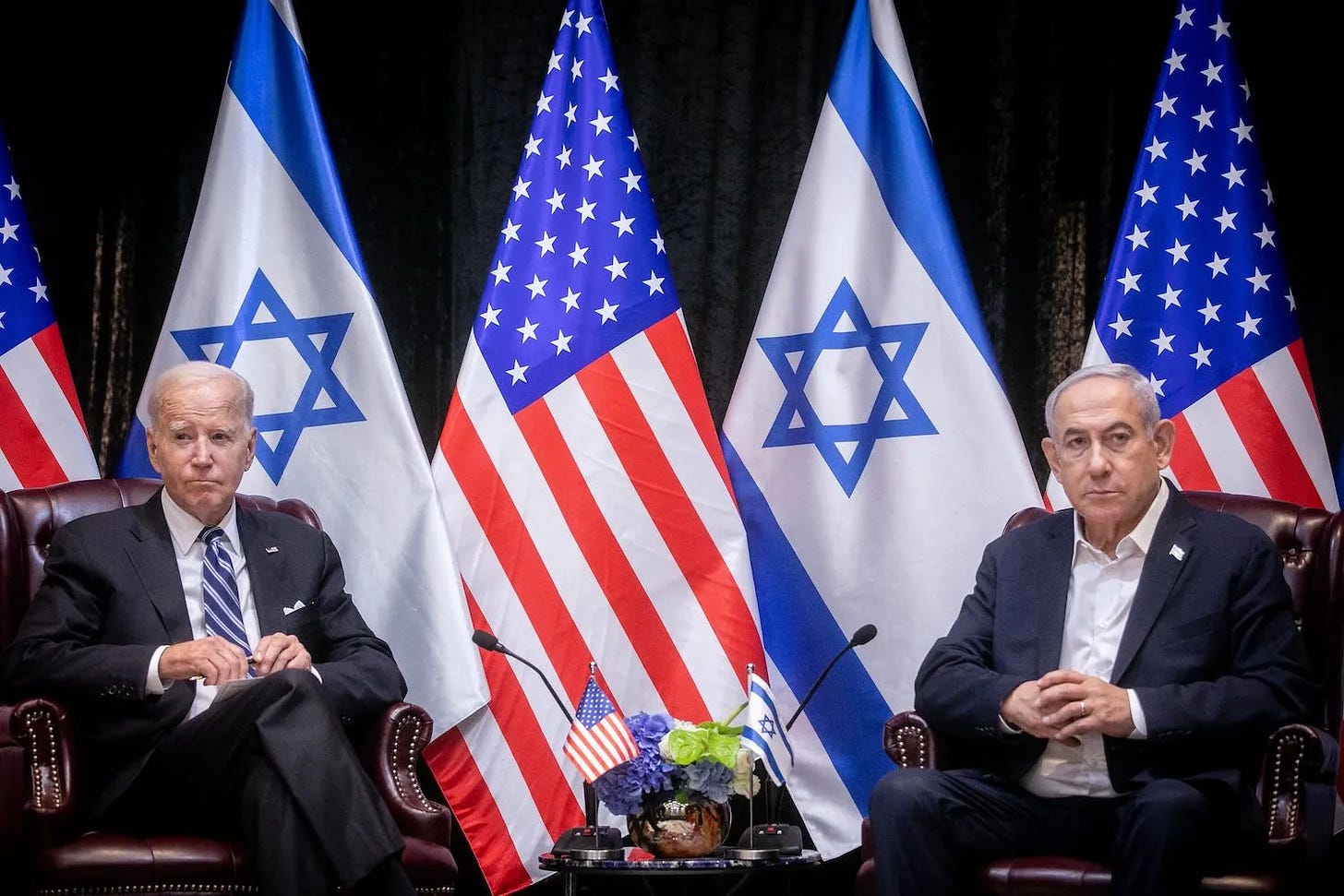 U.S. President Joe Biden (left) and Israeli Prime Minister Benjamin Netanyahu sit beside each other in a identical leather-upholstered chairs. Israeli and American flags hang from poles behind the men, and a table between them holds flowers, smaller flags, and two microphones pointed at either leader.
