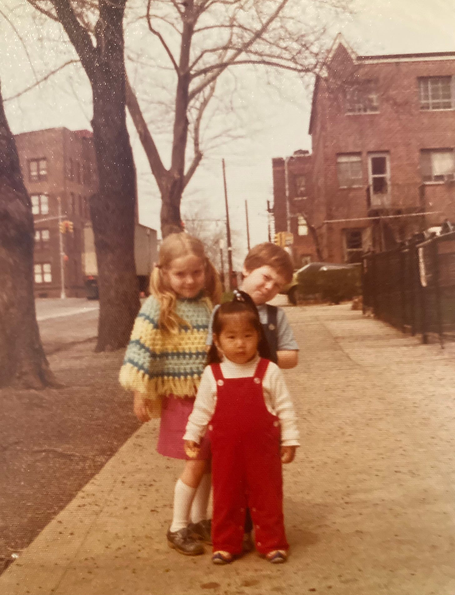 picture of me as a toddler in Queens, NYC on a street on what looks like an early spring or late fall day in red overalls and pigtails with brick buildings in background. two children with fair hair and wearing 70s style clothing are behind me, posing for the photograph. i do not know who they are.