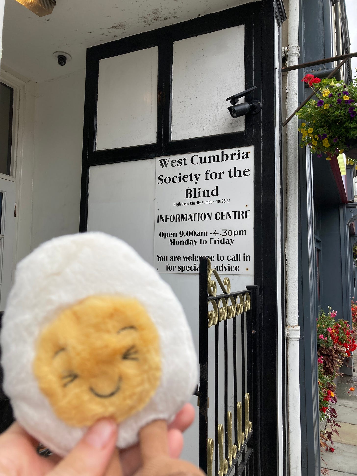 Photograph of a soft toy boiled egg being held in front of a sign that reads 'West Cumbria Society for the Blind' Information Centre. It is a small building on a high street with metal gates.