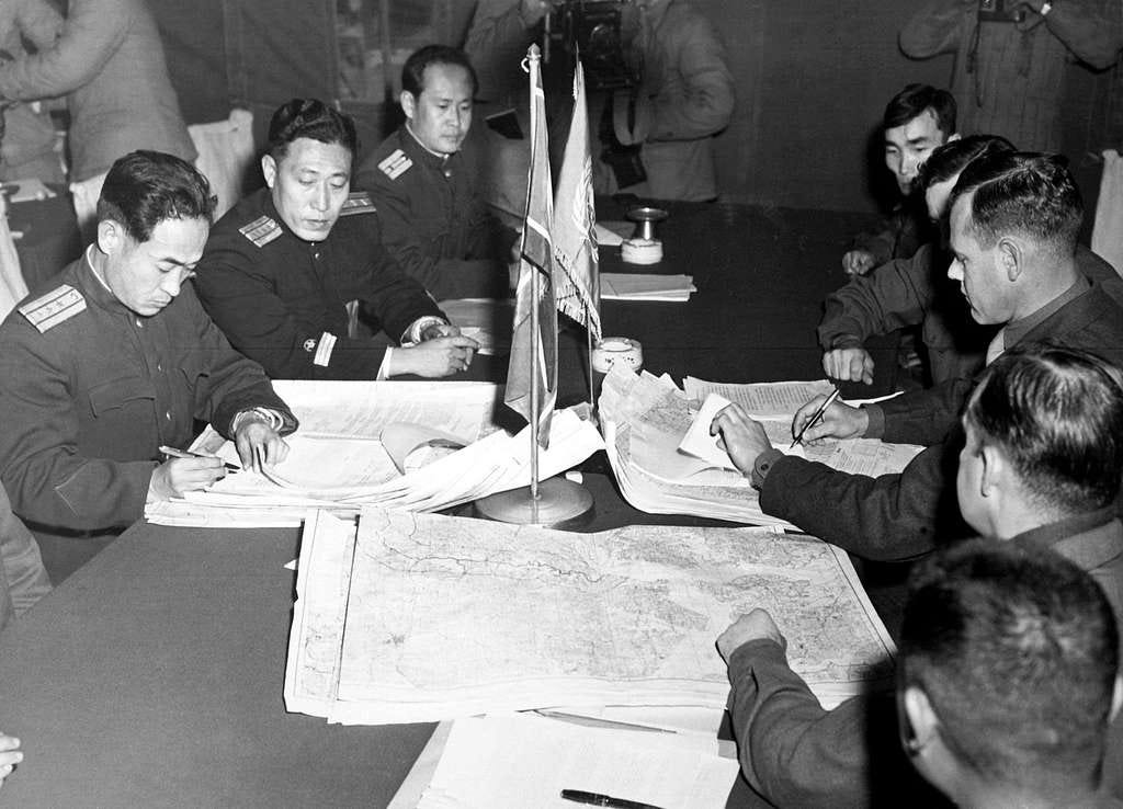 Black and white photo of a negotiating table between North Korean, United States and United Nations officials at the Panmunjon cease fire talks. There are three North Korean officers on the left side of the table facing the camera with 5 US and UN officials on the right side of the table with the sides and backs of their heads captured in the image. The officials are in formal uniforms. On the table are maps of the lines of demarcation the officials are validating with signatures. Black and white photo of a negotiating table between North Korean, United States and United Nations officials at the Panmunjon cease fire talks. There are three North Korean officers on the left side of the table facing the camera with 5 US and UN officials on the right side of the table with the sides and backs of their heads captured in the image. The officials are in formal uniforms. On the table are maps of the lines of demarcation the officials are validating with signatures.