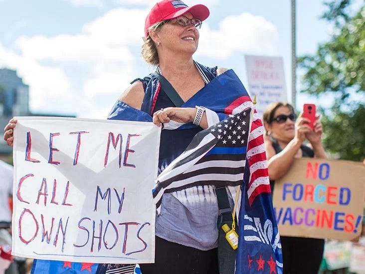 A smiling woman in a red cap and draped in US Flag regalia holds a sign: "Let Me Call My Own Shots" at an outdoor protest. Another sign reads: "No Forced Vaccines!" A smiling woman in a red cap and draped in US Flag regalia holds a sign: "Let Me Call My Own Shots" at an outdoor protest. Another sign reads: "No Forced Vaccines!"