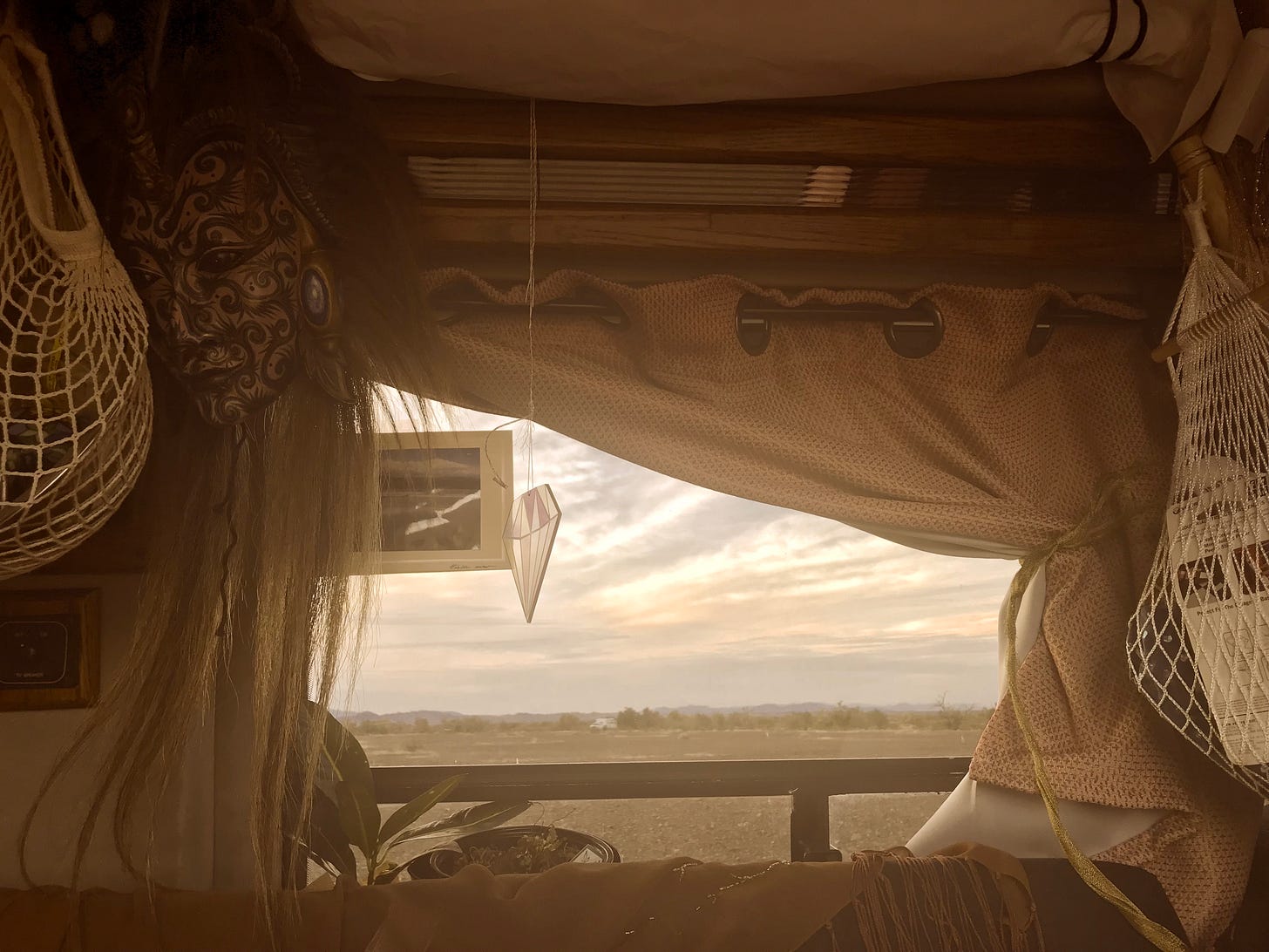 A view of sunrise out the window of a van; inside you can see a mask from Bali and bags of produce hanging from a rod, the top of a couch, and a plant in the windowsill.