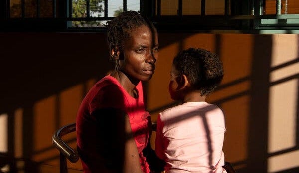 A woman in a red shirt and pink pants sits on a chair with a child in her lap.