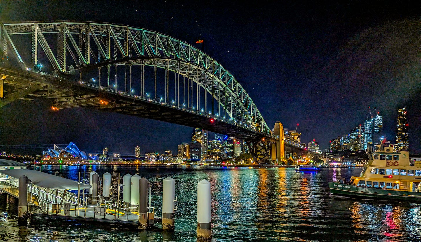 Sydney Harbour Bridge lit up at night, the lights reflecting off of the water as a ferry passes in front. 