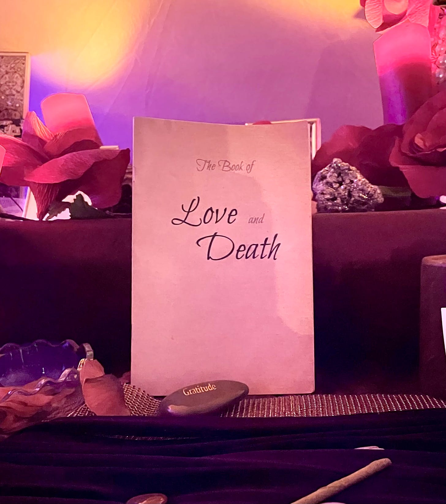 A close-up photo of The Book of Love and Death, standing on an altar table in purple and yellow lights.